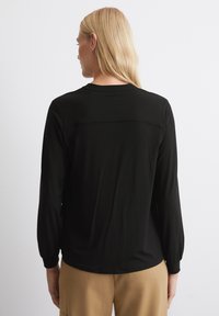 Top nero a maniche lunghe realizzato in tessuto liscio, caratterizzato da un collo rotondo e un delicato dettaglio della cucitura posteriore, abbinato a pantaloni beige.