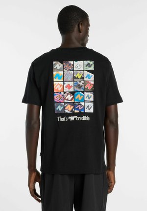 Mann trägt ein schwarzes T-Shirt mit einem bunten Raster aus "N"-Logos und dem Text "That's 'N'credible" auf der Rückseite, steht vor einem weißen Hintergrund.