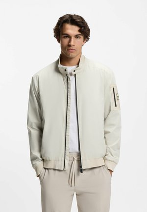 Ung mand iført en lys beige lynlåstrøje med en mærkat på ærmet, hvid skjorte under og beige bukser med snor i taljen.