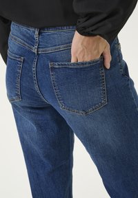 Tamsiai mėlynos džinsinės kelnės su klasikiniu penkių kišenių dizainu, matomais siuvinėjimo detalėmis ir šiek tiek dėvėtu tekstūru.