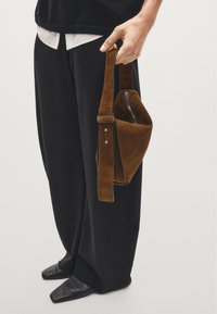 Brown suede bag with a short strap held by a hand; features a zip closure and silver hardware. Black wide-leg trousers and pointed black shoes are visible.