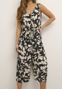 Mono floral en negro y beige con escote en V, diseño sin mangas, cintura elástica y piernas anchas recortadas; tejido ligero con un patrón texturizado.
