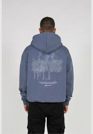 Person, die einen blauen Hoodie mit verzerrter weißer Grafik und Text auf dem Rücken trägt, kombiniert mit schwarzen Cargohosen, steht vor einem einfarbigen Hintergrund.