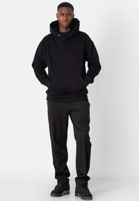Schwarzer Hoodie mit einer Fronttasche, strukturiertem Stoff und flachen Kordeln, kombiniert mit schwarzen Hosen und Sneakers. Schlichtes, modernes Design.