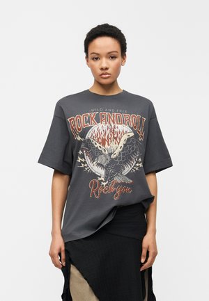Donna che indossa una maglietta oversize grigia scura con la scritta "Rock and Roll" e una grafica di un'aquila davanti, abbinata a una gonna nera a strati.