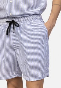 Άτομο που φορά κοντά παντελόνια με μπλε και λευκές ρίγες και μαύρο κορδόνι, με το χέρι στην τσέπη και φαίνεται μέρος από το αντίστοιχο ριγωτό πουκάμισο.