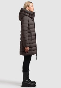 Brązowa kurtka puchowa z wysokim kołnierzem, zamkiem błyskawicznym, kieszeniami bocznymi i kapturem. Kurtka ma pikowany wzór i jest zestawiona z czarnymi legginsami i botkami.