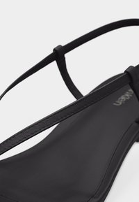 Μαύρο μινιμαλιστικό σανδάλι με λεπτά λουριά που διασταυρώνονται πάνω από το πόδι και επίπεδη σόλα πάνω σε ανοιχτό φόντο.