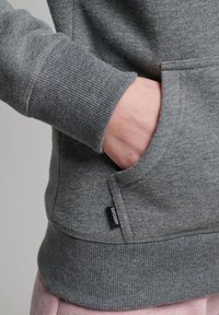 Mano nella tasca frontale di una felpa grigia Superdry con polsini e vita a coste, pantaloni rosa visibili sotto.
