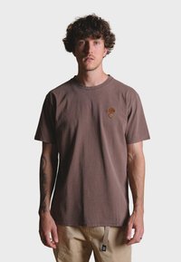 Hnědé bavlněné tri�čko s krátkými rukávy, kulatým výstřihem a malým vyšitým logem stromu na prsou. Model je vyladěn s lehkými kalhotami.