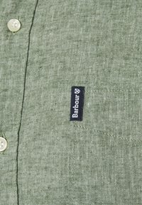 Zelená lněná košile s texturovanou látkou, krémovými knoflíky a černou nášivkou Barbour na levé straně.