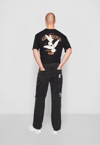 Black cotton t-shirt with back graphic of two white birds, text in orange. Paired with loose black cargo pants with multiple pockets and white shoes.