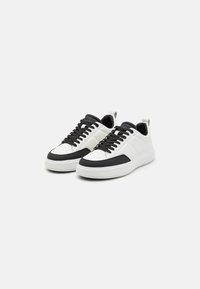 Baskets en cuir blanc et noir avec une finition lisse, bout rond, lacets noirs et semelle en caoutchouc. Présente un tirant au niveau du talon.