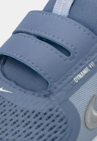 Sportssko i ljusblå mesh med förstärkt tå, dubbla velcroremmar och "DYNAMIC FIT"-branding, med en silverfärgad Nike-logotyp på sidan.