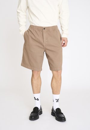 Mand iført beige shorts med elastisk talje, hvide sokker med sort tekst, sorte loafers og en langærmet hvid skjorte, hænderne i lommerne.