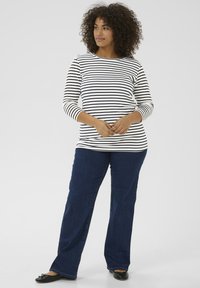 Striped long-sleeve shirt in white and navy, paired with dark blue denim jeans and black flats. Curly hair, standing pose.