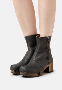 Černé kožené kotníkové boty se zipem na boku, robustním dřevěným podpatkem a drsnou podešví. Vyznačují se hladkým texturem a strukturovaným designem.