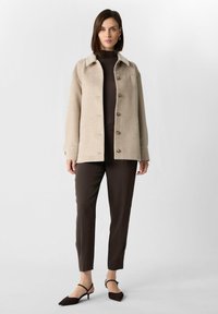Giacca beige in misto lana con colletto classico, bottoni frontali e due tasche laterali, abbinata a pantaloni marroni aderenti e décolleté scuri con cinturino.