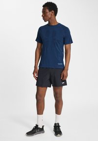 Marinblå atletisk t-shirt med en texturerad logotypdesign, tillsammans med svarta shorts. Modellen har på sig svarta sneakers och vita träningsstrumpor.