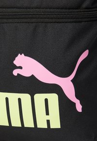 Črna tkanina torba z svetlo rožnatim logotipom Puma in velikim, svetlo zelenim besedilom. Površina prikazuje teksturiran, trpežen material.