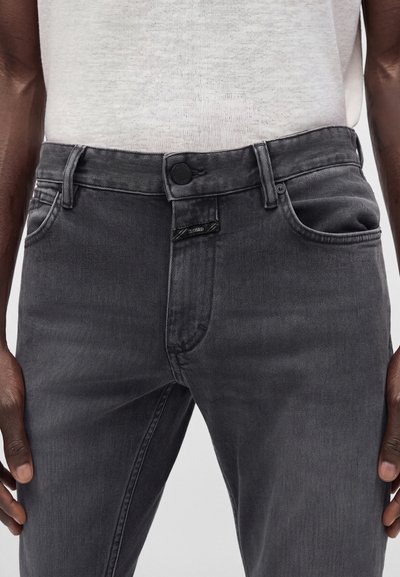 Homme portant un jean en denim noir délavé et un t-shirt blanc semi-transparent, les mains reposant sur les côtés.