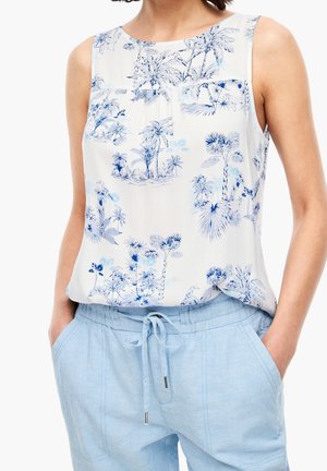 Blouse blanche sans manches avec imprimé tropical bleu associée à un pantalon bleu clair à cordon, mains dans les poches, portée par une personne debout.