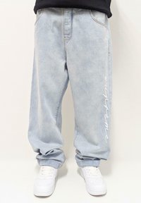 Gaiši zili džinsa baggy bikses ar nelielu izbalējumu, ar baltu izšūtu "Supreme" tekstu uz kreisā kājas un standarta kabatām.