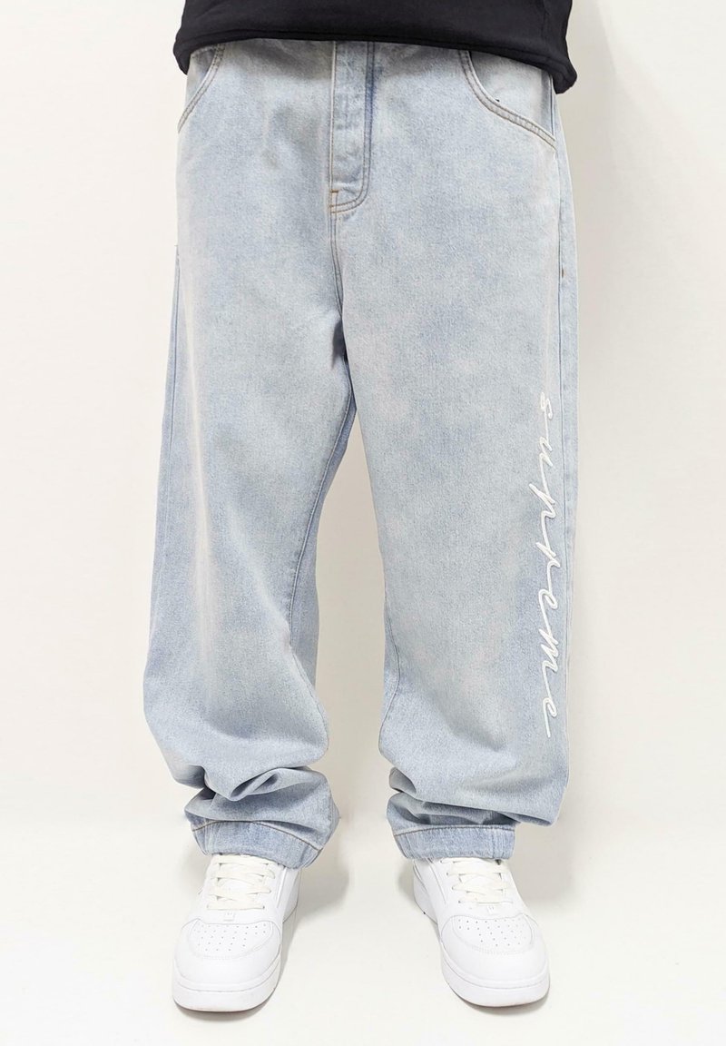 Gaiši zili džinsa baggy bikses ar nelielu izbalējumu, ar baltu izšūtu "Supreme" tekstu uz kreisā kājas un standarta kabatām.