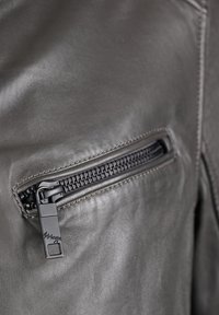 Szara skórzana kurtka z kieszenią na zamek błyskawiczny, wyposażoną w teksturowany zamek i czarny uchwyt, podkreślająca gładkie i matowe powierzchnie tkaniny.