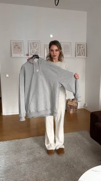 Sweatshirt cinza com capuz, apresentando um bolso frontal e um pequeno logotipo no peito. O modelo está segurando-o pelo cabide. Textura macia, corte oversized.