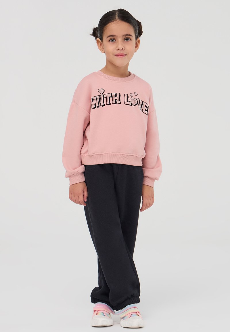 Felpa rosa con la scritta "WITH LOVE" in lettere nere, abbinata a pantaloni della tuta grigio scuro con polsini elasticizzati. Sneakers bianche con lacci colorati.