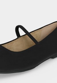 Chaussure plate Mary Jane noire avec une fine bride unique et une doublure intérieure beige visible sur un fond clair uni.