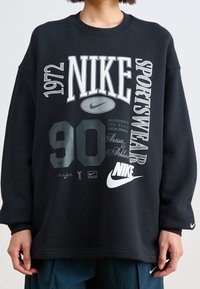 Person in einem schwarzen Nike-Sweatshirt mit mehreren Logos und Texten, darunter "1972", "Nike", "Sportswear" und "90" in unterschiedlichen Schriftarten und Größen.
