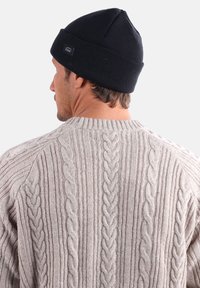 Bonnet en tricot noir avec un sommet pointu, porté sur un pull en tricot grisé à torsades présentant des motifs en relief et un col côtelé.