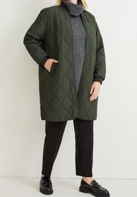 Quilted green long jacket with a zip front, side pockets, and ribbed cuffs, layered over a gray turtleneck and black trousers. Black loafers.