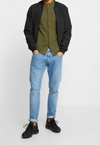 Zwarte bomberjack over een olijfgroene overhemden met knopen, lichtblauwe spijkerbroek en zwarte sneakers. Casual outfit, standaard manchetten, geen zichtbare patronen.