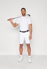 Vit pikétröja med korta ärmar och ett svart bälte, kombinerat med vita shorts. Figuren håller en golfklubba över axeln.