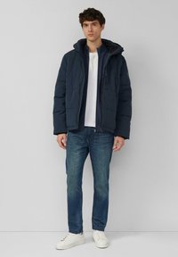 Marineblauwe gewatteerde jas met capuchon, ritssluiting aan de voorkant en geribbelde manchetten, gedragen over een wit shirt en blauwe spijkerbroek, gecombineerd met witte sneakers.