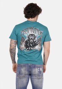 T-shirt in cotone turchese con design grafico di un pantera nera che ruggisce, arricchito da testo ed elementi geometrici in bianco e blu.