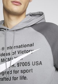 Grå huvtröja med mörkgrå detaljer, med svart text och en vit Nike-swoosh. Mjuk tyg med dragsnören vid huvan.