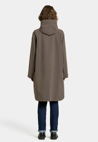 Long manteau à capuche de couleur marron foncé, en matériau imperméable. Présente une coupe droite, des fermetures à pression et des manches longues. Associé à un jean foncé et des bottes.