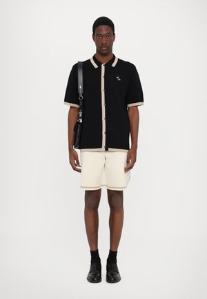 Schwarzes Strickshirt mit beigem Besatz, kurze Ärmel, Knopfverschluss. Cremefarbene Shorts mit beigen Akzenten. Schwarze Schuhe und eine Schultertasche ergänzen das Outfit.