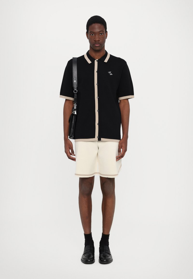 Schwarzes Strickshirt mit beigem Besatz, kurze Ärmel, Knopfverschluss. Cremefarbene Shorts mit beigen Akzenten. Schwarze Schuhe und eine Schultertasche ergänzen das Outfit.