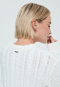 Pull en tricot blanc avec un motif en relief et une coupe décontractée. Poignets côtelés et une petite étiquette noire à l'arrière du col. Matériel doux et texturé.