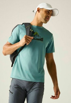 Hombre con gorra blanca, camiseta con gráfico de montaña en color azul verdoso, pantalones grises y lleva una mochila negra colgada de un hombro.