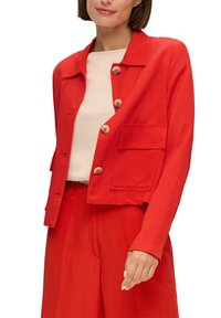 Veste rouge courte en tissu léger, avec un col pointu, de grands boutons et des poches latérales pour plus de détail.