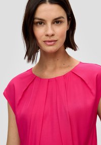 Blusa rosa brillante con maniche corte, davanti plissettato e scollatura rotonda. Tessuto liscio, leggero e senza motivi o accenti visibili.