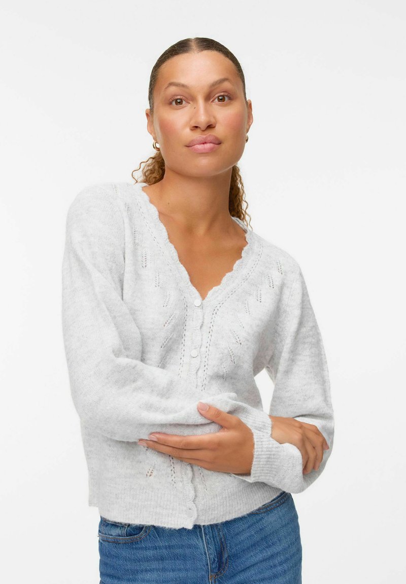 Cardigan en tricot gris avec un col en V festonné, fermeture par boutons à l'avant et motifs décoratifs. Texture douce et manches ajustées.