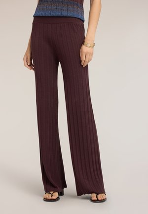 Femme portant un pantalon large côtelé marron foncé taille haute, associé à des sandales noires à bout ouvert et plusieurs bracelets dorés au poignet.