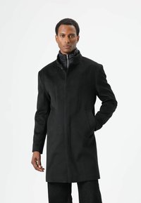 Zwarte jas met een hoge kraag, gemaakt van zacht materiaal. Het heeft zijzakken en een strakke, getailleerde pasvorm, gedragen over een donker binnenjasje.